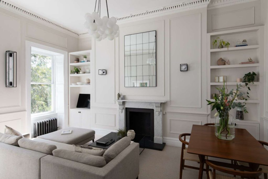 A room with cream-colored walls, shelves on wall filled with books, table with a glass jar plant, chairs under the table