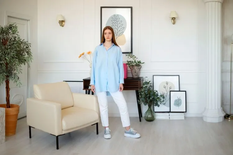 young woman in blue shirt, and white pants stands in a stylish living room with plants, artwork, and a beige armchair