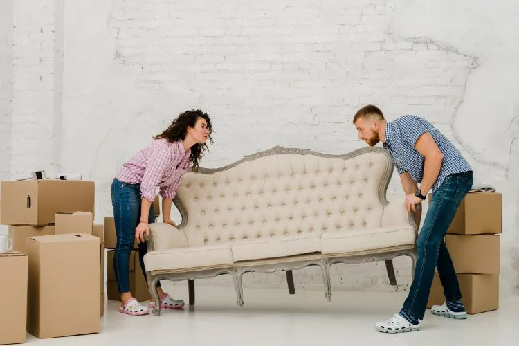 man and woman lift a vintage beige sofa in a room with white brick walls, surrounded by moving boxes